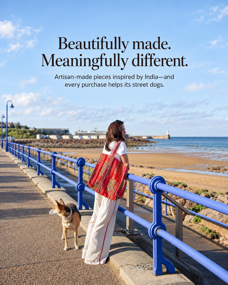A woman stands on a seaside promenade by a blue railing, wearing a bright red and gold patterned tote bag. A dog is beside her, with the ocean, rocks, and a pier in the background.
