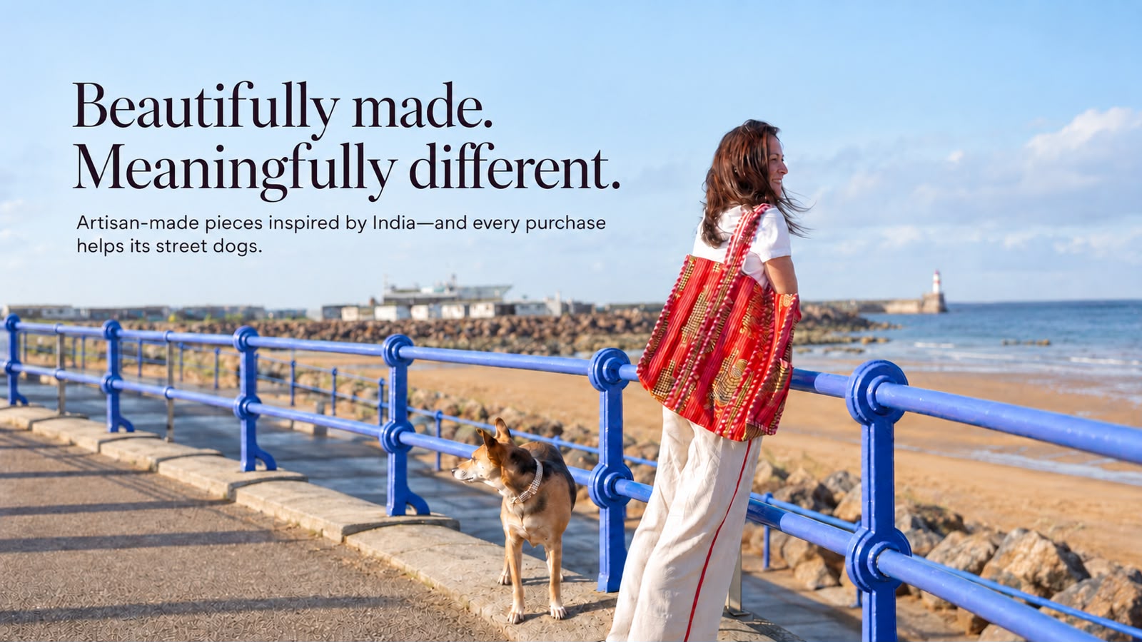 A woman stands on a seaside promenade by a blue railing, wearing a bright red and gold patterned tote bag. A dog is beside her, with the ocean, rocks, and a pier in the background.